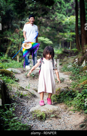 Japanese girl wearing sun rose pale robe et sac à dos de transport debout dans une forêt, l'homme dans l'arrière-plan. Banque D'Images