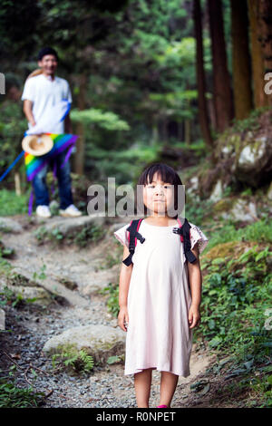 Japanese girl wearing sun rose pale robe et sac à dos de transport debout dans une forêt, l'homme dans l'arrière-plan. Banque D'Images