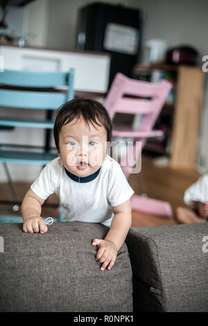 Portrait d'enfant japonais debout sur un canapé gris, à la caméra à curieusement. Banque D'Images