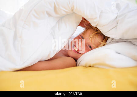 Smiling boy au lit de se cacher sous une couette Banque D'Images