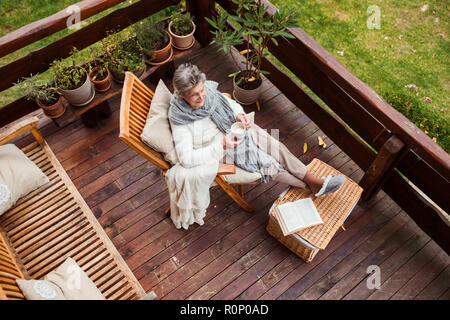 Une vue de dessus d'une femme âgée avec un livre à l'extérieur sur une terrasse en automne. Banque D'Images