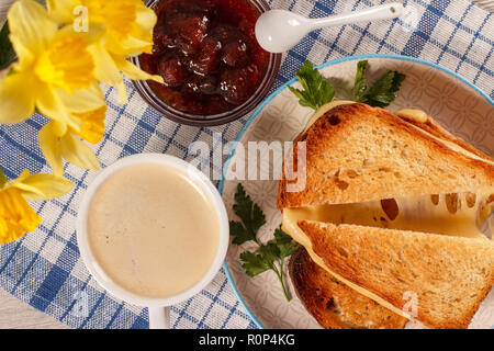 Tranches de pain grillé avec du fromage et persil vert sur plaque blanche, tasse de café, bol en verre avec de la confiture de fraise et cuillère, jonquilles jaune, bleu Banque D'Images