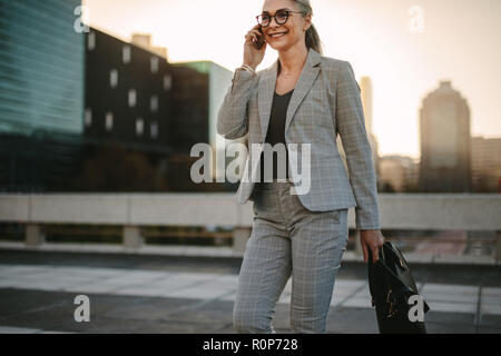 Belle femme senior marchant à l'extérieur sur la parlant au téléphone. Businesswoman walking on city street avec téléphone mobile. Banque D'Images