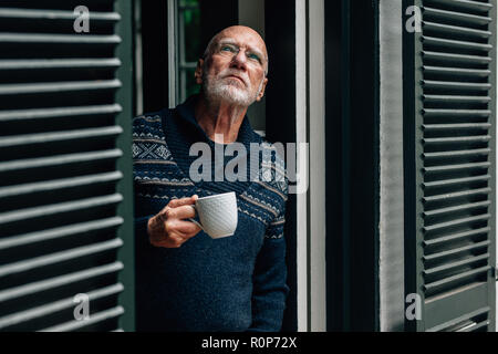 Old man looking up debout à la fenêtre avec une tasse de café dans la main. Man savourer une tasse de café debout à la fenêtre à la maison. Banque D'Images