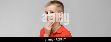 Portrait de petite blonde happy smiling caucasian boy in tee-shirt orange avec la main près de son visage sur fond gris Banque D'Images