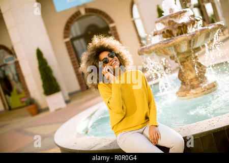 Aftican american jeune femme est assise près de la fontaine et les pourparlers sur le smartphone. Banque D'Images