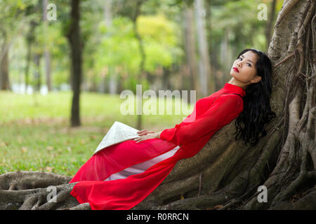 Bien mince jeune Vietnamien dame habillé en vêtements traditionnels rouge détente sur les racines aériennes dans un parc public, couchée sur son dos alors qu'elle était stare Banque D'Images