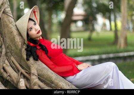 Magnifique jeune femme vietnamienne vêtue d'un costume rouge traditionnel à rêver qu'elle se penche en arrière confortablement contre le tronc d'un arbre en milieu urbain pa Banque D'Images