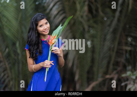 Smiling Young Vietnamese Woman in Blue Dress Holding Fleurs fraîches avec des grandes feuilles vertes en regardant la caméra. Banque D'Images