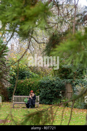 Femme portant un chapeau rouge assis sur un banc en bois dans un jardin ou parc dans la saison d'automne à la cathédrale de Chichester. Banque D'Images