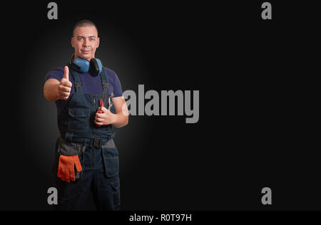 Homme forte et confiante en bleu mécanicien permanent overal orange avec des gants, casque blanc, la protection des oreilles et des outils sur un fond noir Banque D'Images