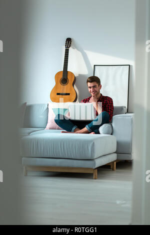 Les jeunes positifs millénaire man sitting on sofa in living room with laptop seul sur week-end à la maison Banque D'Images
