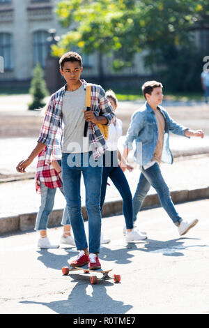 Beau jeune écolier américain africain équitation skateboard avec ses camarades de l'arrière-plan flou sur marche Banque D'Images