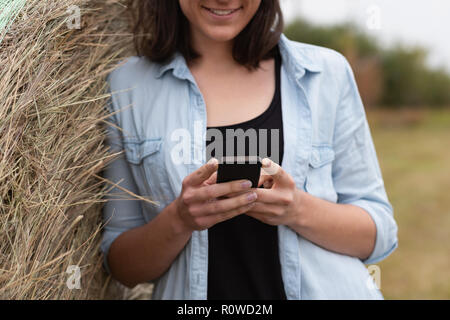 Woman using mobile phone while leaning on hay bale Banque D'Images