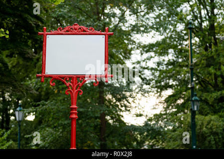 Cadre métallique décoratif rouge avec bannière blanche pour le texte. Dans l'arrière-plan les arbres avec des feuilles vertes. Banque D'Images