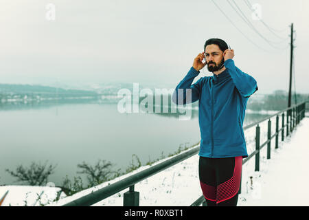 Un homme coureur avec le casque sur ses oreilles prenant une pause dans l'espace public au cours de la formation d'hiver à l'extérieur à côté de la rivière. Copier l'espace. Banque D'Images