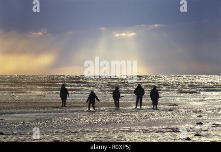 Cinq personnes marchent sur la plage à marée basse et regardent les rayons du soleil et de la lumière crépusculaire brillent d'un ciel orageux. La mer et les sables brillent et les gens sont en silhouette Banque D'Images