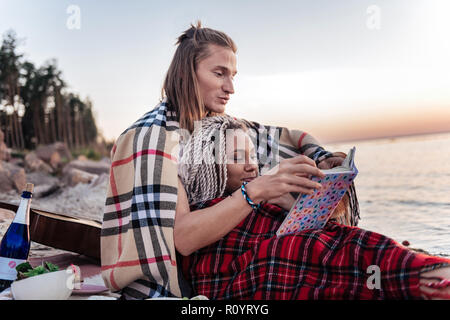 Cute couple aimant avec des plaids sur les épaules lecture livre après pique-nique Banque D'Images