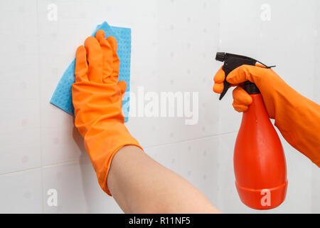 Les mains dans les gants en caoutchouc orange détient bouteille de détergent et de serviette en microfibre bleu. Femme est lave-mur avec du carrelage blanc dans la salle de bains. Cle Banque D'Images