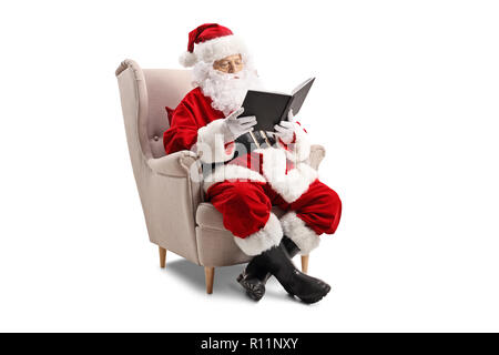 Père Noël assis dans un fauteuil et de lire un livre isolé sur fond blanc Banque D'Images