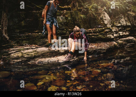 Deux femmes les randonneurs par un étang d'eau en montagne. Femme assise sur rocher et de mettre sa main dans l'eau avec l'ami par et à la recherche. Banque D'Images