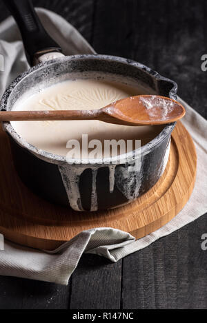 Lait bouilli dans une casserole noire et débordé sur les bords, d'une cuillère de bois, sur une planche à découper, sur une table rustique. Lumière faible image. Banque D'Images