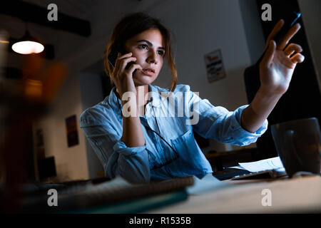 Image of young woman designer graphique à l'aide de pc ordinateur parler par téléphone mobile travaillant de nuit dans le bureau. Banque D'Images