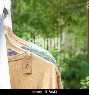 Vêtements biologiques. T-shirts de couleur naturel pendaison sur cintres en bois dans une rangée. Tag Eco textile. La forêt verte, la nature en arrière plan. Banque D'Images