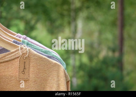 Vêtements biologiques. T-shirts de couleur naturel pendaison sur cintres en bois dans une rangée. Tag Eco textile. La forêt verte, la nature en arrière plan. Banque D'Images