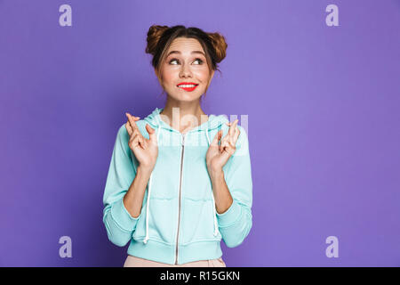 Portrait de jeune fille drôle avec deux petits pains en gardant les doigts croisés et de rêver pour la bonne chance sur fond violet isolé en studio Banque D'Images