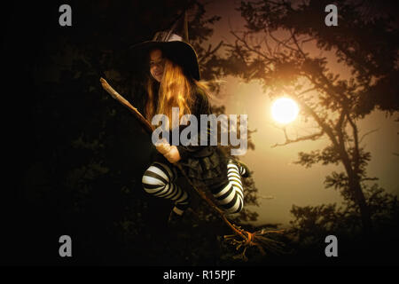 Une petite fille en costume traditionnel d'une sorcière médiévale et un chapeau pointu est voler sur un balai à travers une forêt sombre Banque D'Images