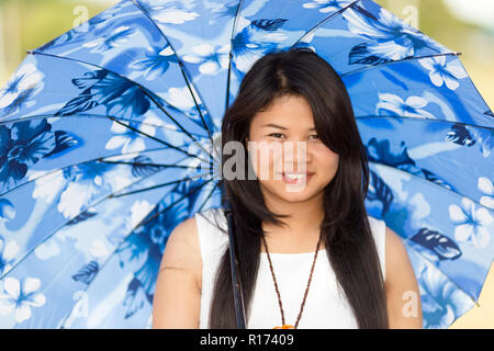 Belle jeune fille thaïe sous un parasol ou parapluie bleu pour la protéger contre le chaud soleil d'été souriant joyeusement à l'appareil photo Banque D'Images