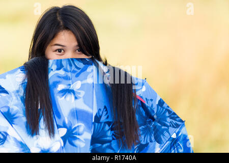 Les jeunes ludique Thai girl peeking sur le haut de son bleu ou un pare-soleil avec un regard amusé alors qu'elle joue à l'extérieur dans la lumière du soleil chaude, avec l'exemplaire Banque D'Images