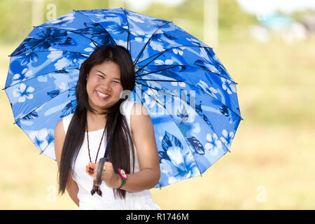 Belle jeune fille thaïe sous un parasol ou parapluie bleu pour la protéger contre le chaud soleil d'été souriant joyeusement à l'appareil photo Banque D'Images