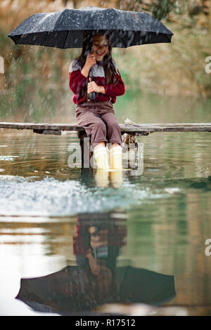 Fille enfant avec parapluie siège au pont de bois et laughes sur fond de rivière dans la pluie. Banque D'Images