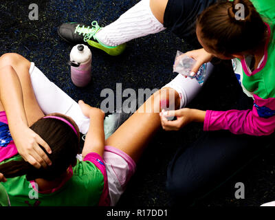 Un gardien de fille avec un nettoyage des ongles peints d'un genou blessé coéquipier avec de l'eau au cours d'un match de football sur un terrain en gazon synthétique. Banque D'Images