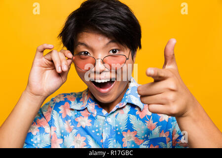 Close up portrait of a choqué asian man in sunglasses sur fond jaune isolé, looking at camera, montre de l'index Banque D'Images