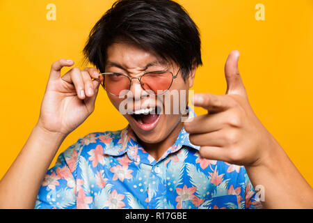 Close up portrait of a choqué asian man in sunglasses sur fond jaune isolé, looking at camera, doigt, clignant de l'oeil Banque D'Images