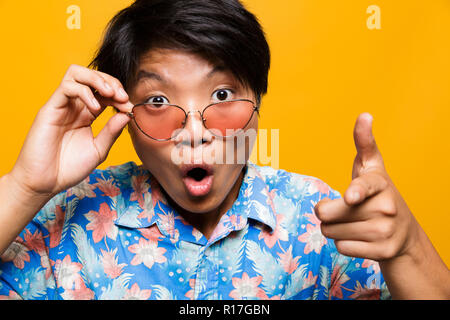 Close up portrait of a choqué asian man in sunglasses sur fond jaune isolé, looking at camera, montre de l'index Banque D'Images