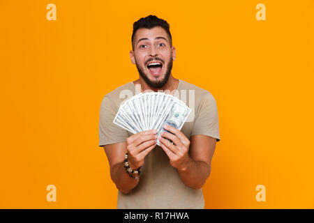 Portrait d'un jeune homme barbu excité en t-shirt sur isolé sur fond orange, montrant les billets d'argent Banque D'Images