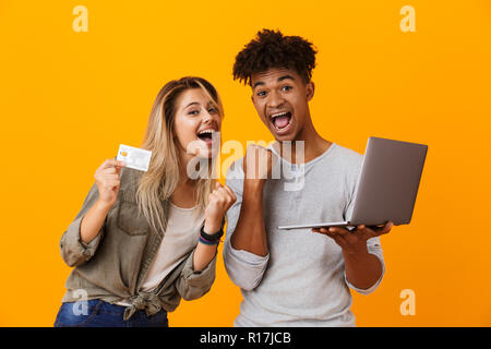 Photo de heureux cheerful young couple aimant isolés sur fond jaune à l'aide d'un ordinateur portable faire de geste gagnant holding credit card. Banque D'Images