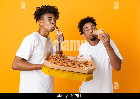 Deux hommes heureux amis africains dans t-shirts isolés sur fond jaune, manger pizza savoureuse d'un fort Banque D'Images