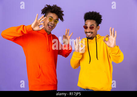 Deux heureux homme africain amis vêtus de sweats à capuche et lunettes colorées isolé sur fond violet permanent, montrant le geste ok Banque D'Images