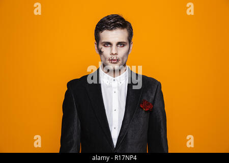 Photo de zombie effrayant halloween sur l'homme portant costume classique et maquillage creepy looking at camera isolated over fond jaune Banque D'Images