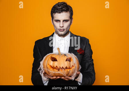 Photo de scary halloween homme mort sur le port de costume classique et maquillage creepy holding carved pumpkin sur fond jaune isolé Banque D'Images