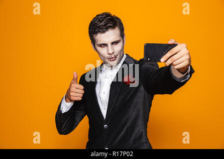 Photo de scary halloween homme mort sur le port de costume classique et creepy en tenant un miroir sur téléphone mobile selfies sur fond jaune isolé Banque D'Images