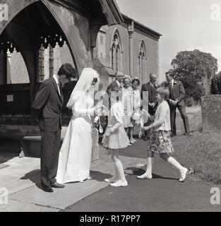 1967, historique, un mariage d'été et de deux jeunes filles présente des cuillères en bois pour la mariée le marié avec commandes à l'extérieur de l'église, England, UK. Une cuillère en bois pour l'épouse, c'est un cadeau traditionnel de bonne chance et de bibelots datant de nombreuses années le symbole de la femme pour être une 'bonne' Cook pour son nouveau mari. Banque D'Images