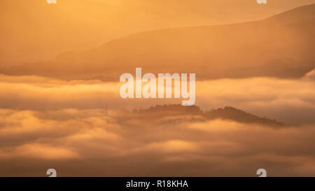 Soleil lumineux lumière dorée sur un misty Valley dans le Peak District, England, UK. Une colline arborée se lève comme une île au-dessus du brouillard. Banque D'Images