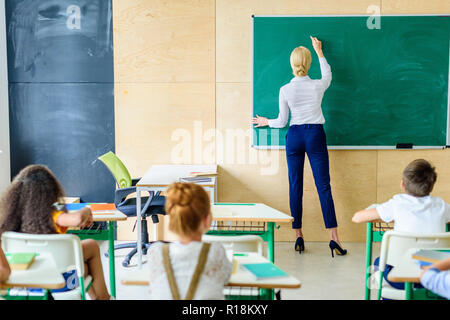 Vue arrière d'élèves à l'enseignant au pendant qu'elle écrit on chalkboard Banque D'Images