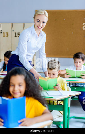 Smiling teacher contrôle la façon dont les enfants lire des livres pendant la leçon Banque D'Images